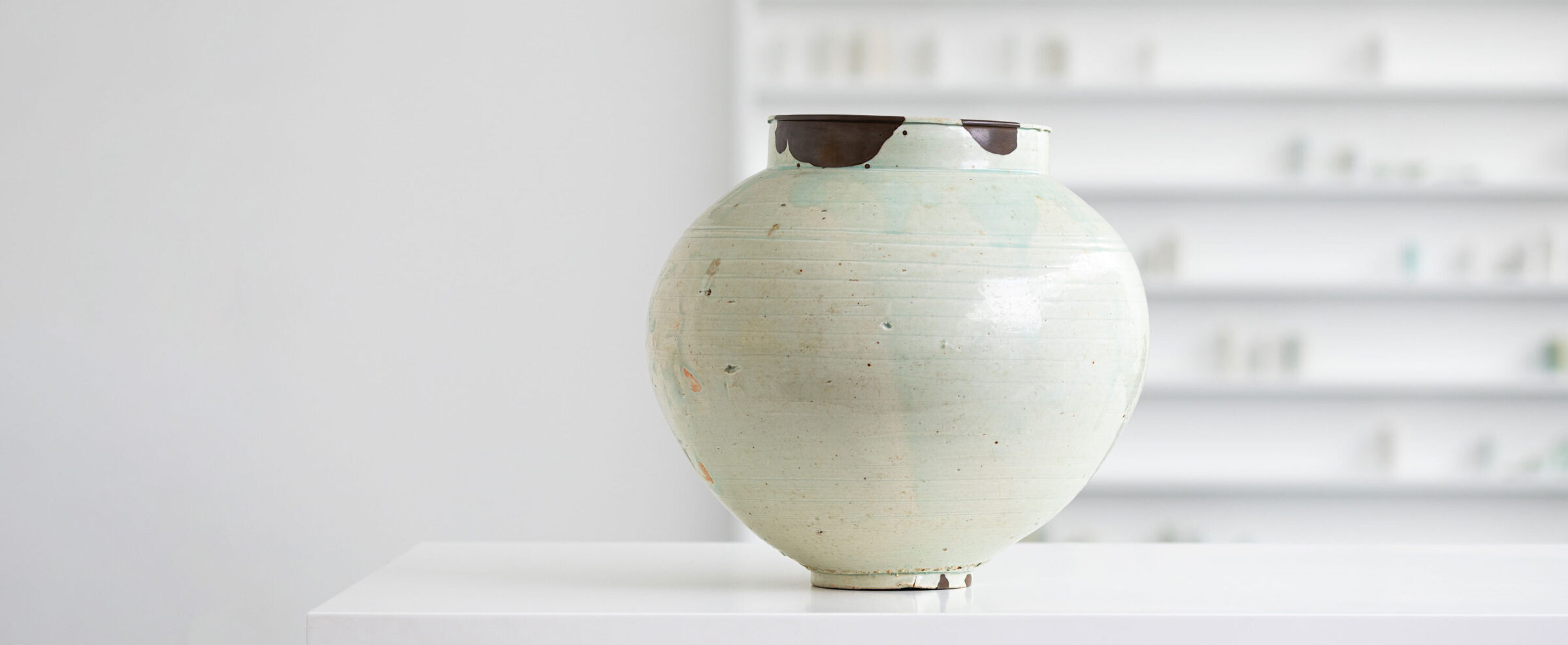 A round porcelain jar, glazed in white with subtle blue-green tones sits on a white plinth. In the background, and slightly out of focus, is an installation of small white porcelain vessels within a white vitrine.