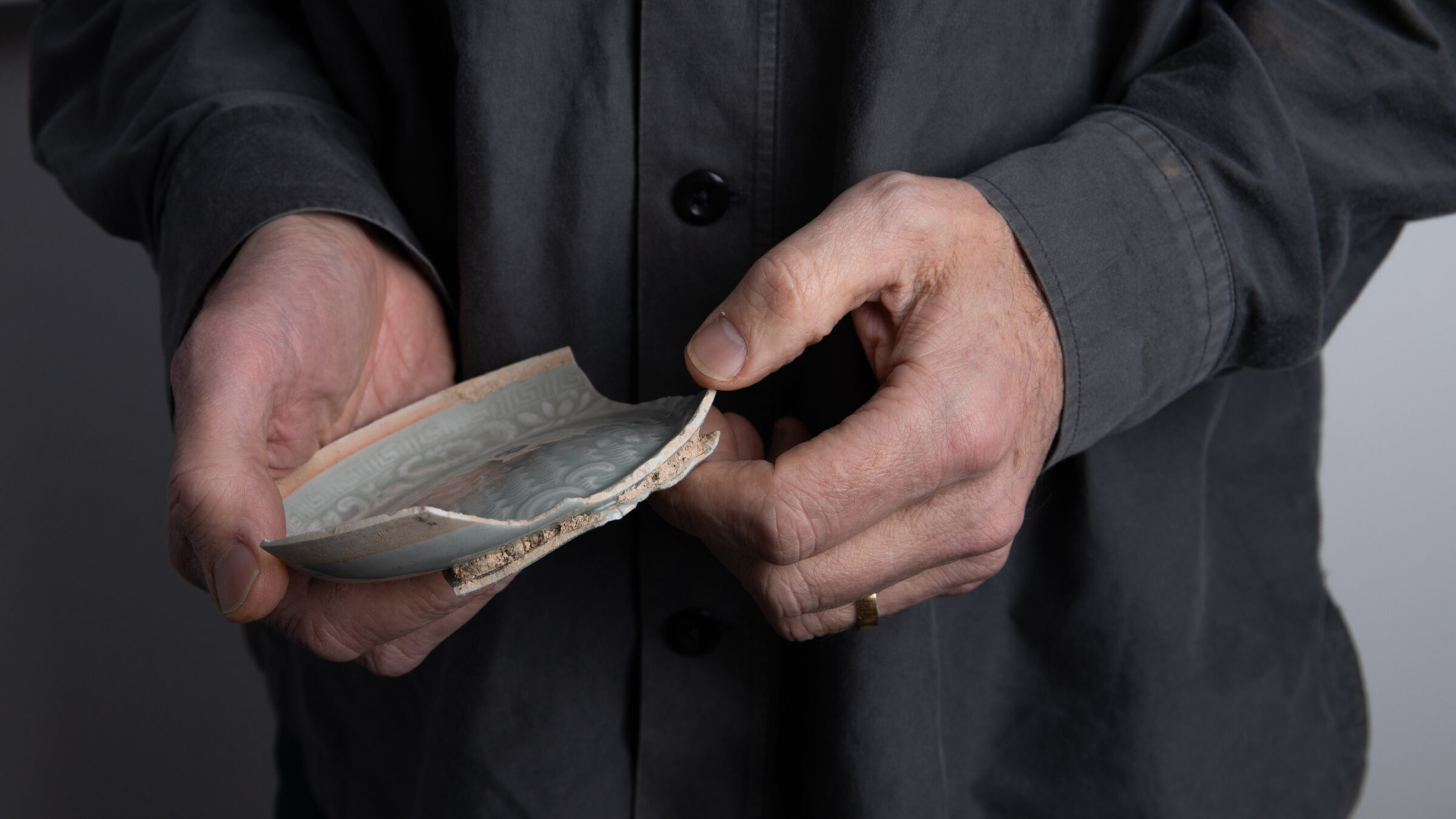 A close up image of Edmund de Waal's hands holding a broken shard of pottery. The shard is a shallow dish with a pale green glaze, the edges are rough and jagged.
